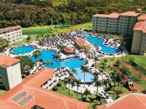 Vista aérea: resort, edifícios, piscinas azuis, palmeiras, espreguiçadeiras, quadras esportivas, vegetação.