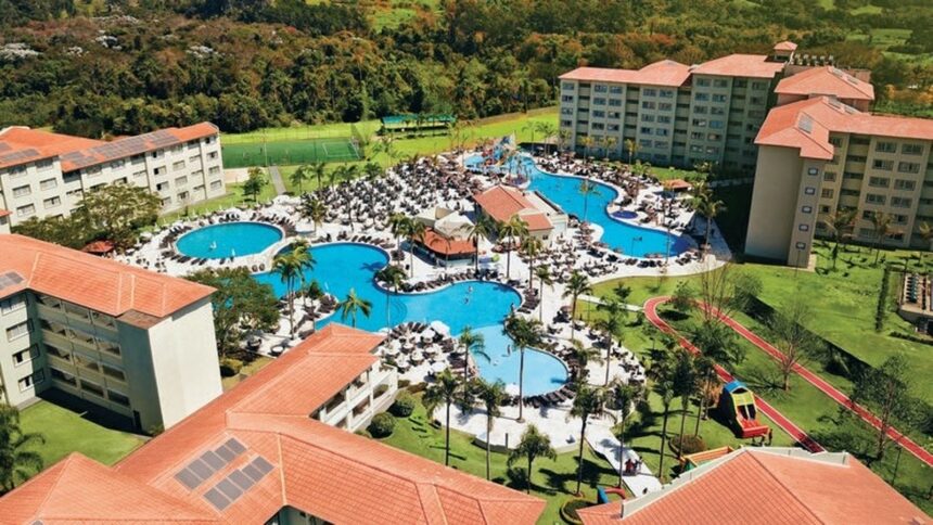Vista aérea: resort, edifícios, piscinas azuis, palmeiras, espreguiçadeiras, quadras esportivas, vegetação.