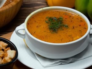 Tigela branca de caldo alaranjado com tempero verde, acompanhada de croutons e pão.