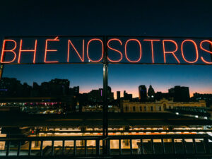 Placa de neon vermelho 'BHÉ NOSOTROS' com skyline da cidade ao anoitecer.