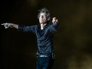Mick Jagger no palco, segurando microfone, vestindo camisa de bolinhas, apontando