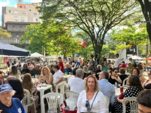 Multidão de pessoas sentadas em mesas ao ar livre sob árvores, com prédios ao fundo, em evento diurno.