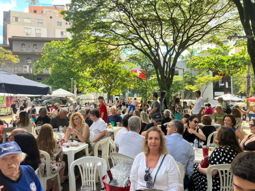 Multidão de pessoas sentadas em mesas ao ar livre sob árvores, com prédios ao fundo, em evento diurno.