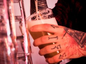Mão tatuada segura copo com cerveja loira sendo servida em ambiente de bar