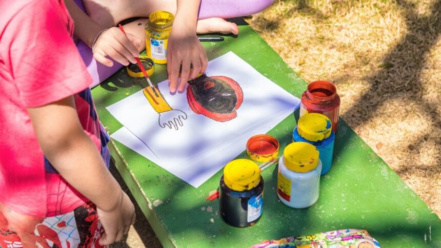 Mãos infantis pintando desenho de rosto com tintas coloridas sobre uma mesa verde.
