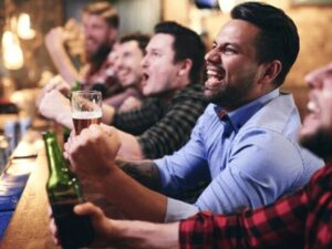 Grupo de homens animados com cerveja, torcendo e celebrando em um bar.