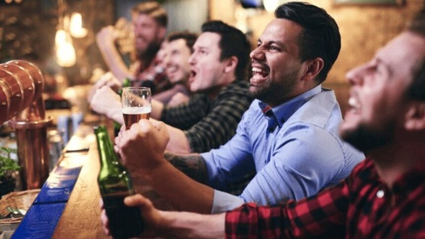 Grupo de homens animados com cerveja, torcendo e celebrando em um bar.