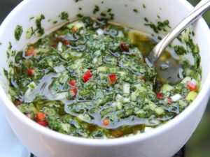 Tigela branca com chimichurri fresco, ervas picadas, pimenta vermelha, óleo e uma colher de metal
