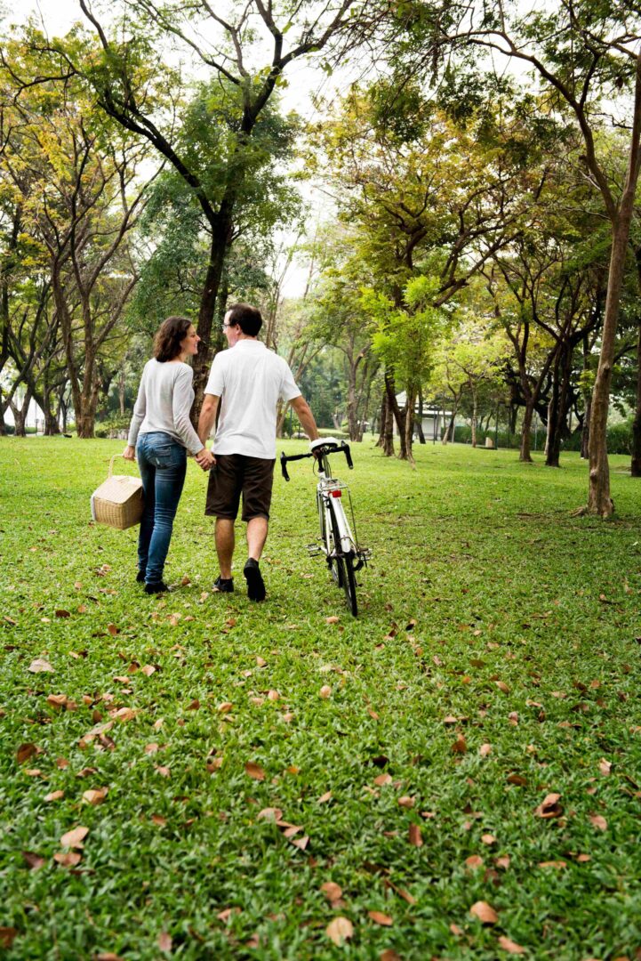 Um casal caminha de mãos dadas em um parque com bicicleta e cesta de piquenique