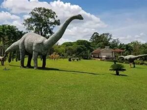 Três estátuas de dinossauros em um parque verde com céu azul e uma casa ao fundo
