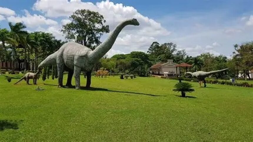 Três estátuas de dinossauros em um parque verde com céu azul e uma casa ao fundo