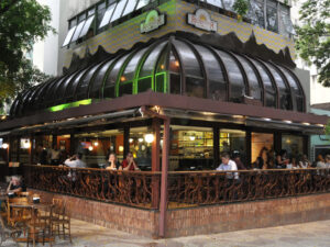 Pessoas sentadas em bar-restaurante com mesas externas, balcão interno e telhado de vidro curvo