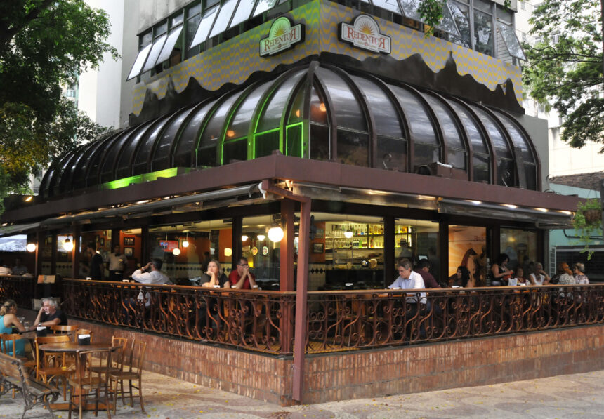 Pessoas sentadas em bar-restaurante com mesas externas, balcão interno e telhado de vidro curvo
