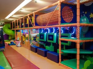 Parque infantil indoor colorido com estrutura de escalada, escorregador verde, túnel e videogames