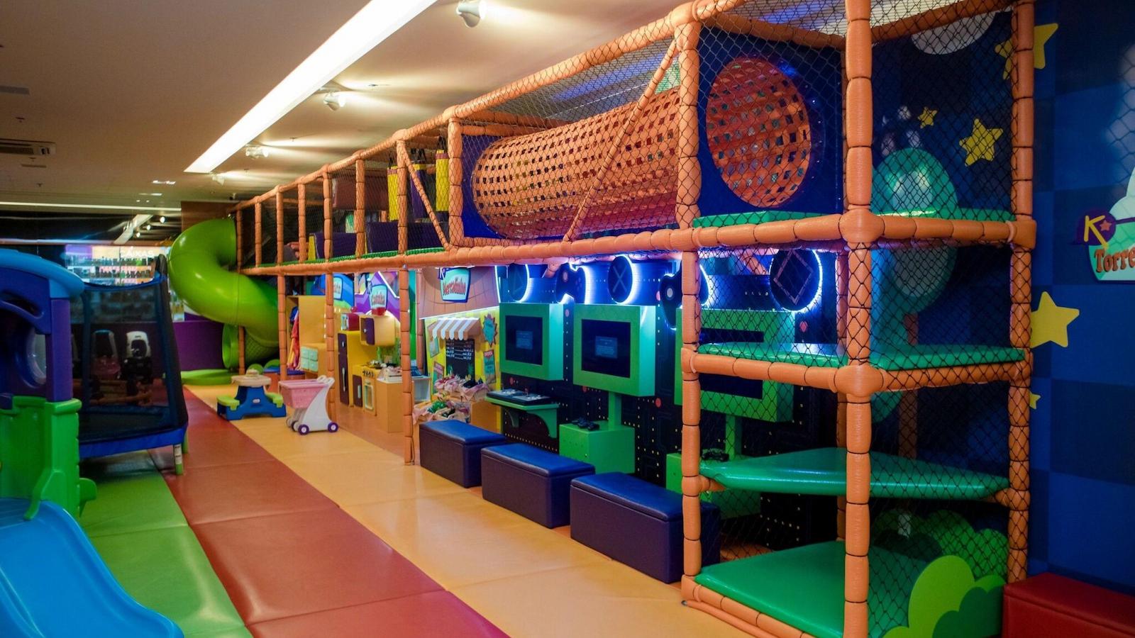 Parque infantil indoor colorido com estrutura de escalada, escorregador verde, túnel e videogames