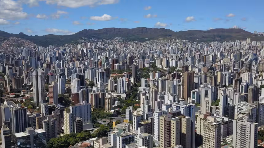 Vista aérea panorâmica de uma BH com arranha-céus e montanhas ao fundo sob céu azul.