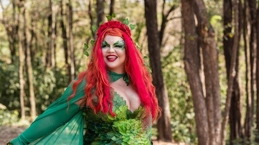 Mulher fantasiada de Hera Venenosa com cabelo vermelho, maquiagem verde, sorrindo em parque arborizado