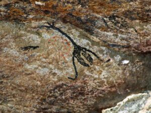 Desenho preto estilizado de figura humana ou animal em parede de rocha com tons marrons e vermelhos