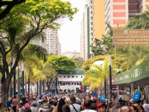 Multidão em festa de rua com árvores, prédios e placas, incluindo "MINASCENTRO"
