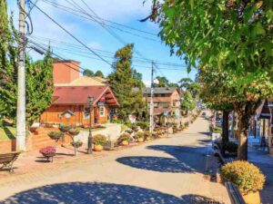 Rua pavimentada com casas de madeira, árvores, bancos e vasos de flores em uma cidade serrana ensolarada