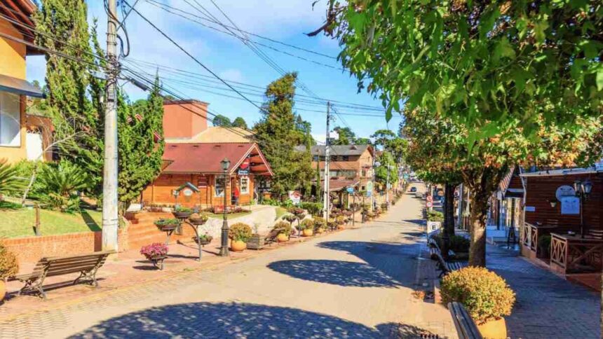 Rua pavimentada com casas de madeira, árvores, bancos e vasos de flores em uma cidade serrana ensolarada
