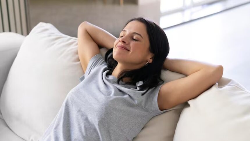 Mulher sorrindo, olhos fechados, relaxando no sofá com as mãos atrás da cabeça