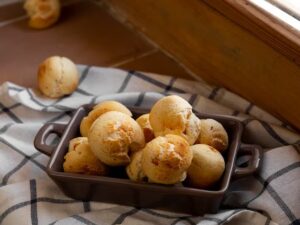 Pão de queijo dourado em travessa sobre pano xadrez ao lado de janela iluminada