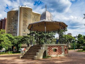 Coreto de metal e tijolos em parque urbano, com árvores frondosas e edifícios altos