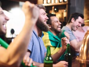 Homens em bar assistindo a um jogo de futebol, comemorando com cervejas e bandeira brasileira