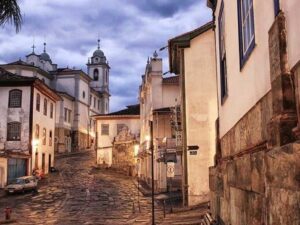 Rua de paralelepípedos com casarões coloniais, igreja e carro estacionado, iluminada por postes