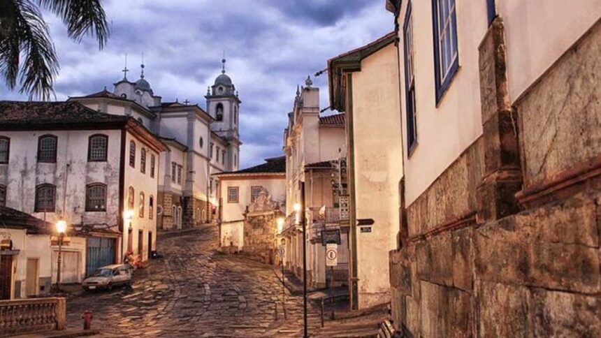 Rua de paralelepípedos com casarões coloniais, igreja e carro estacionado, iluminada por postes