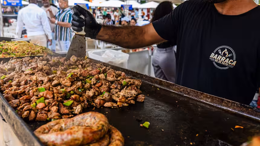 Cozinheiro de camiseta preta com logo "BARRACA DO ZÉ NETO" mexe carne na chapa