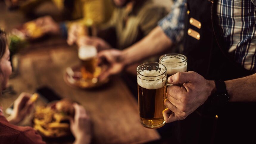 Garçom segurando duas canecas de cerveja artesanal em bar com clientes desfocados ao fundo
