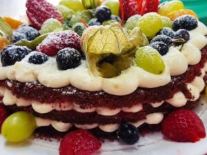 Bolo de camadas com massa escura e creme branco, decorado com frutas frescas e açúcar de confeiteiro