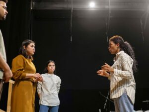 Quatro jovens adultos em palco escuro, uma mulher gesticula enquanto outros observam