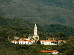 Igreja branca com torre pontiaguda e edifícios em uma paisagem montanhosa densamente arborizada