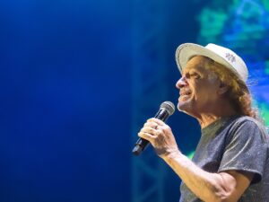 Alceu Valença, cabelos longos e chapéu branco, cantando em um microfone em palco azul