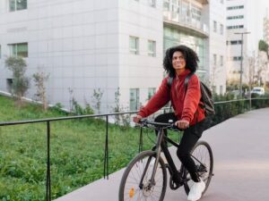 Mulher jovem com mochila e casaco vermelho pedala bicicleta preta em caminho urbano