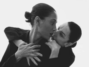 Duas mulheres em preto e branco com rostos próximos, uma com cabelo preso e anel no dedo