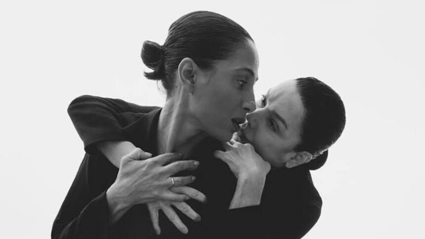 Duas mulheres em preto e branco com rostos próximos, uma com cabelo preso e anel no dedo