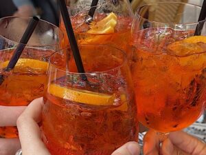 Mãos seguram quatro copos de Aperol Spritz alaranjados com gelo, laranja e canudos pretos