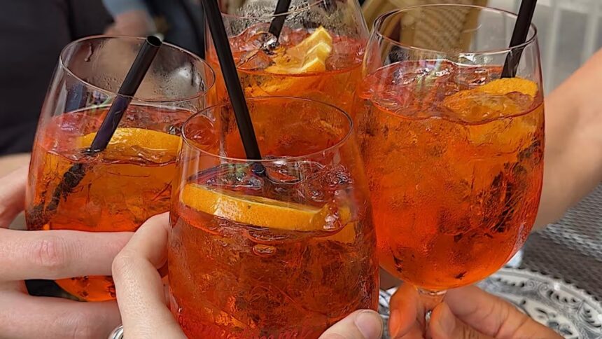 Mãos seguram quatro copos de Aperol Spritz alaranjados com gelo, laranja e canudos pretos