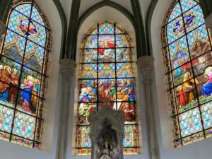 Três vitrais coloridos detalhados em igreja, com cenas bíblicas e estátua no altar central