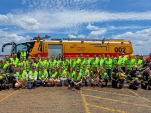 Grupo de pessoas, algumas de coletes e bombeiros, posam em frente a caminhão BH Airport