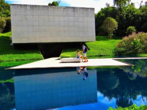 Dois visitantes em plataforma refletidos em água, edifício concreto e gramado verde