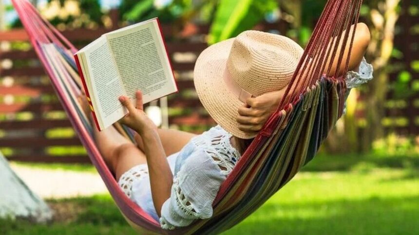 Pessoa de chapéu de palha e roupa branca lendo livro em rede colorida ao ar livre