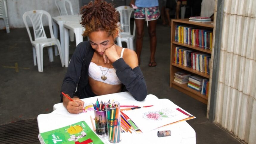 Jovem sorrindo sentada colorindo à mesa com lápis de cor e livros, estante ao fundo