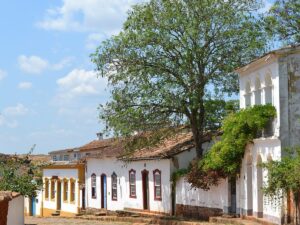 Rua de pedra, casas coloniais brancas, telhados cerâmicos, grande árvore, céu azul claro