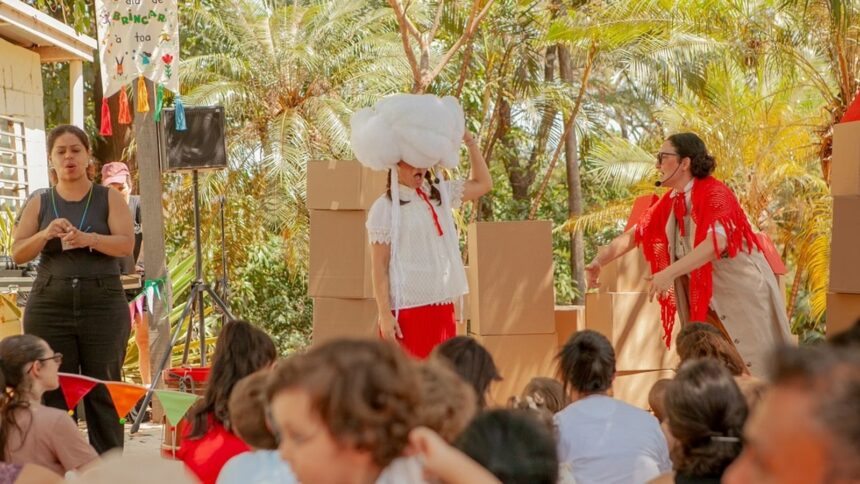 Três mulheres encenam para crianças sentadas em um evento ao ar livre com cenário de caixas.