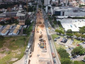 Vista aérea de avenida em obras com maquinário pesado, tráfego de veículos e edifícios ao redor